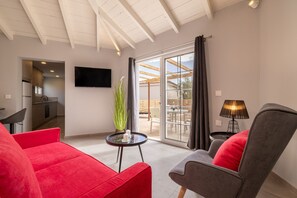 Ein modernes Wohnzimmer mit einem roten Sofa, einem grauen Sessel, einem kleinen Tisch mit einer Kerze, einem an der Wand befestigten Fernseher und einer Glastür zum Außenbereich.