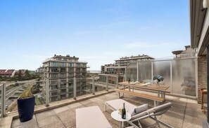 Una terraza en la azotea con barandilla de vidrio, una mesa con sillas y una planta en maceta.