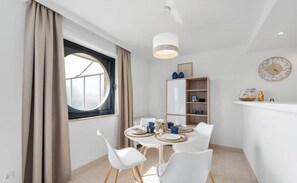 Un área de comedor moderna con una mesa redonda, sillas blancas, una ventana con cortinas, una estantería y un reloj de pared.