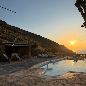 A pool area with lounge chairs, a stone building, and a view of the sunset over the sea. - Sun Holiday Kea – Private Pool & Sea View (Pisses)