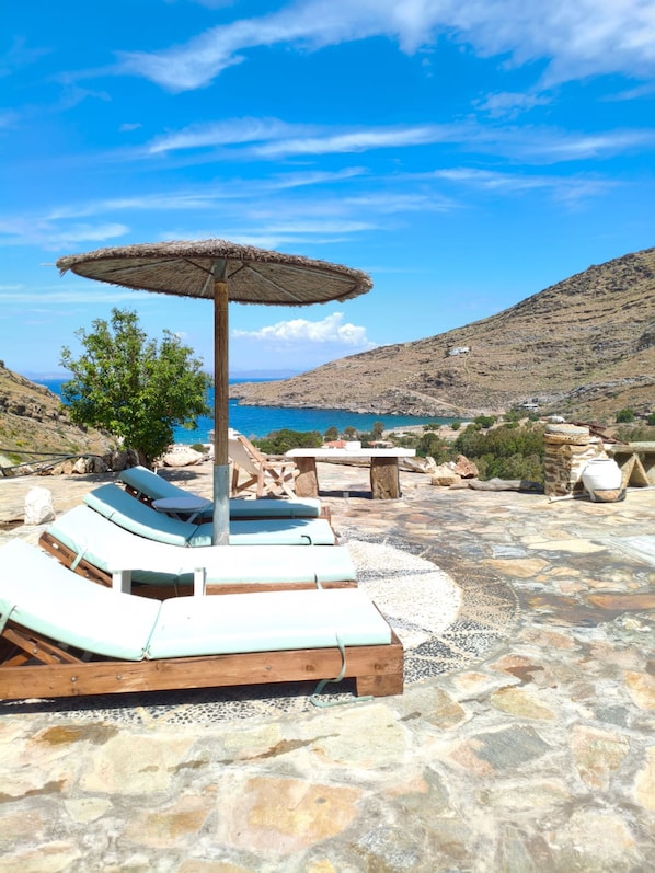A stone patio with lounge chairs and a table, overlooking a body of water and hills. - Sun Holiday Kea – Private Pool & Sea View (Pisses)