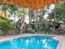 Una piscina en un entorno natural, con árboles y una estructura de techo de paja.
