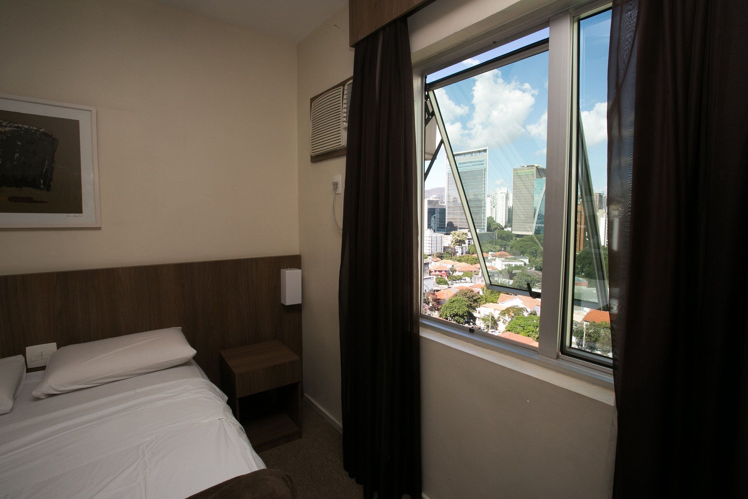 Habitación de hotel con cama, mesita de noche, ventana con cortinas, aire acondicionado de pared y un cuadro enmarcado.