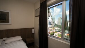 Habitación de hotel con cama, mesita de noche, ventana con cortinas, aire acondicionado de pared y un cuadro enmarcado.