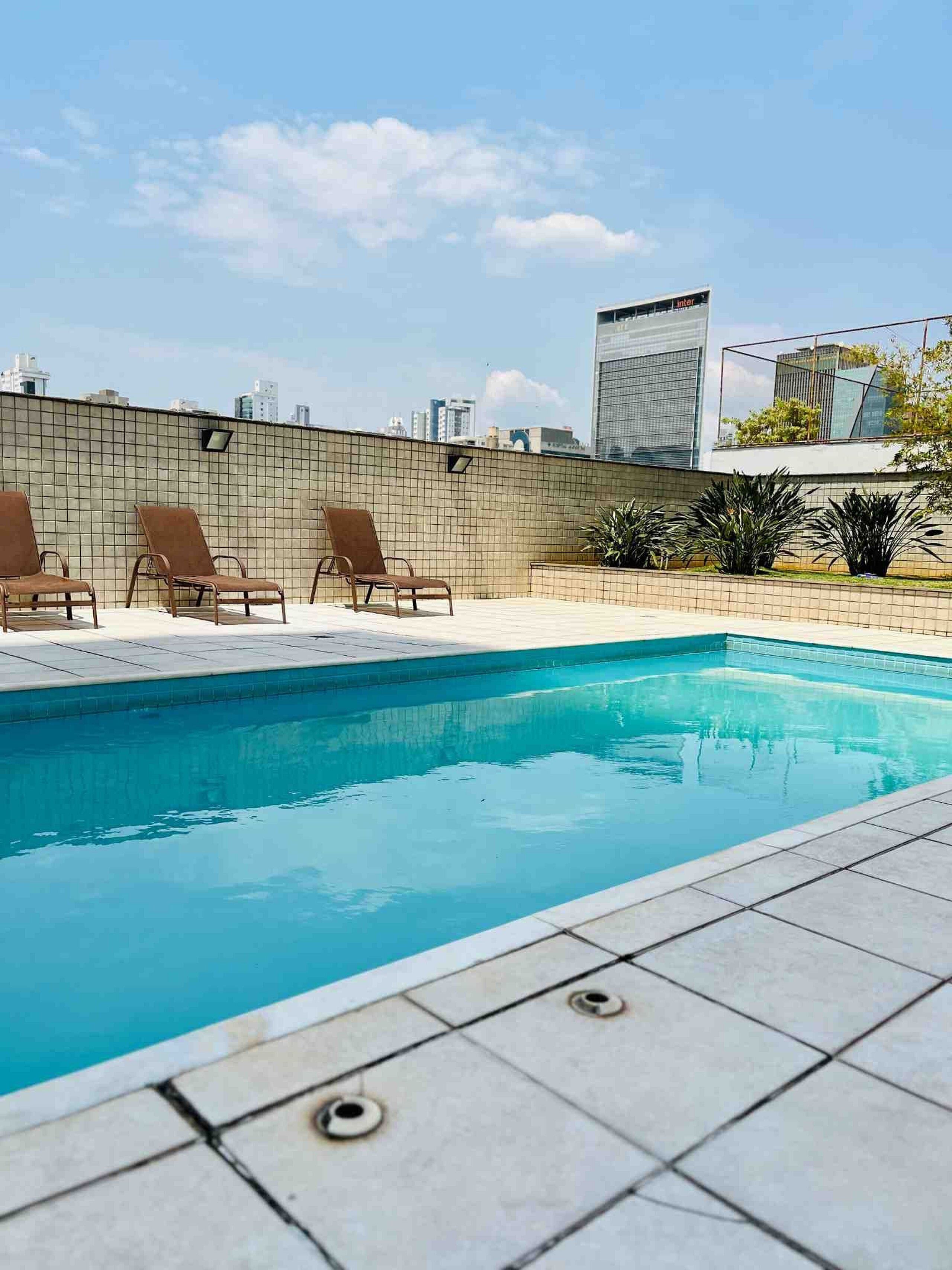 Una piscina en la azotea con tres sillas de descanso, un suelo de baldosas y vistas a la ciudad al fondo.