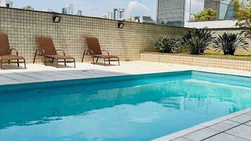 Una piscina en la azotea con tres sillas de descanso, un suelo de baldosas y vistas a la ciudad al fondo.