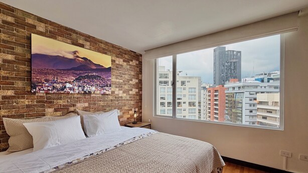 A bedroom with a large bed, a brick wall, a cityscape painting, and a view of high-rise buildings. - Livorno Suites (Quito)