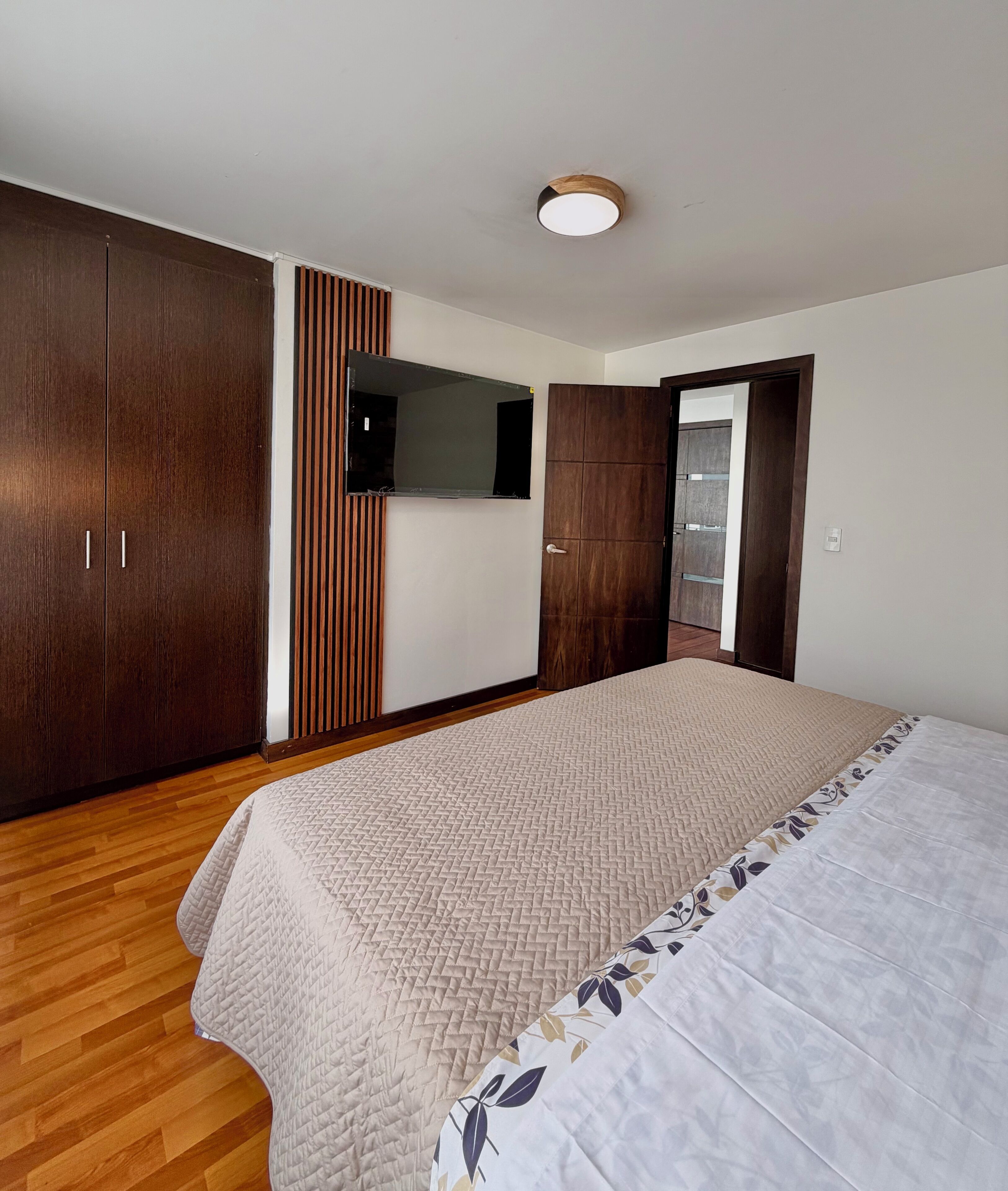 A bedroom with a bed, wooden wardrobe, a television mounted on the wall, and a door leading to another room.