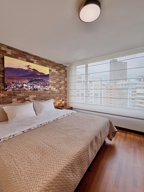 A bedroom with a large bed, a cityscape painting on the wall, a window with a city view, and a brick wall.