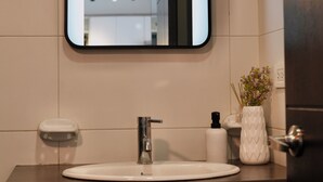 A bathroom with a white sink, a mirror, a towel dispenser, and a vase with flowers. - Livorno Suites (Quito)