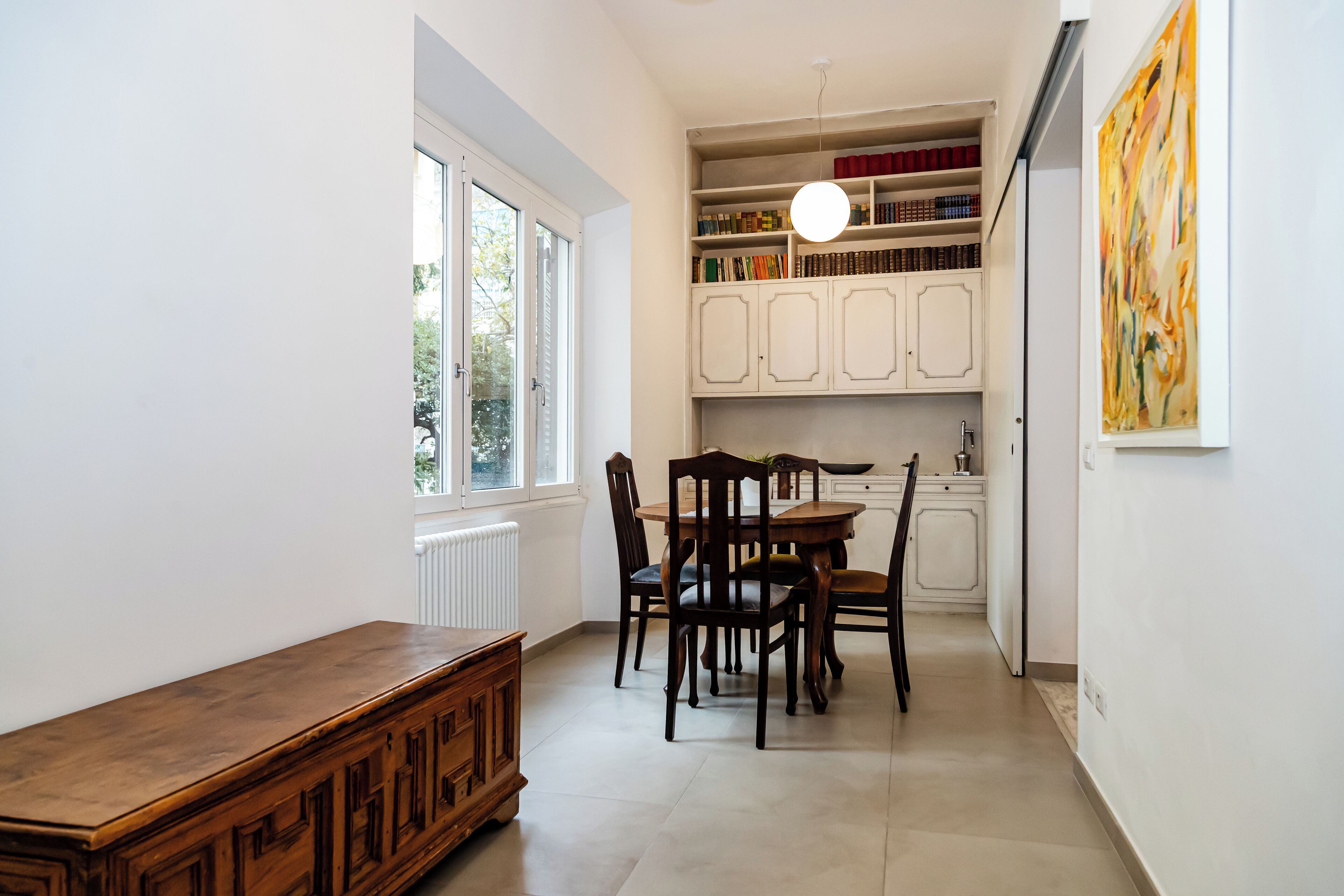 A dining area with a wooden table and chairs, a white cabinet with books, and a window with a view.