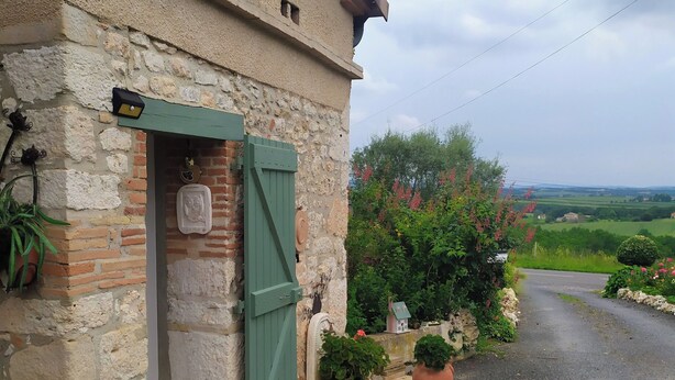 Un bâtiment rustique en pierre, doté d’une porte verte, de plantes en pot et offrant une vue sur la campagne.