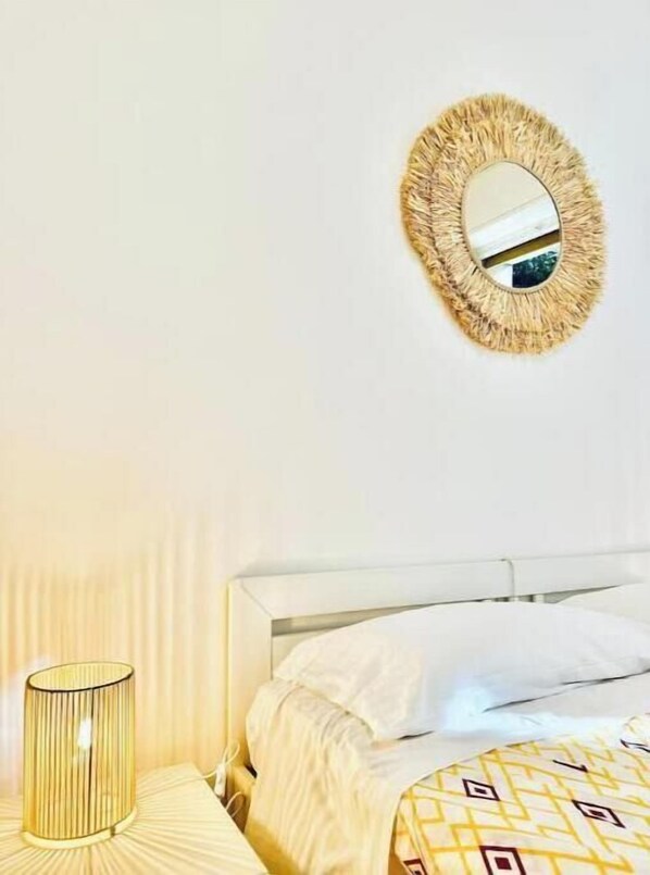 Una cama con ropa blanca, un espejo redondo con marco texturizado y una lámpara cilíndrica sobre una mesita de noche.