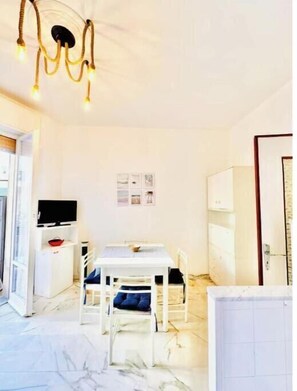 A room with a dining table, chairs, a TV on a stand, a white cabinet, and a door. - Hostdomus - Cozy corner near the sea (Finale Ligure)