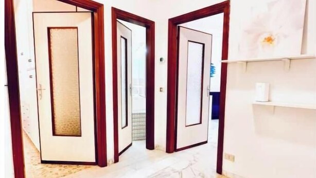 A hallway with three doors, marble flooring, and a white wall with a small shelf. - Hostdomus - Cozy corner near the sea (Finale Ligure)