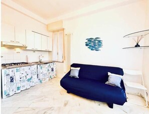 A compact living space with a kitchenette, a dark blue sofa with two pillows, and a wall-mounted fish decoration. - Hostdomus - Cozy corner near the sea (Finale Ligure)