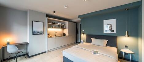 A modern hotel room with a bed, desk, chair, kitchenette, and artwork.