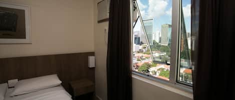 A hotel room with a bed, a bedside table, a window with curtains, a wall-mounted air conditioner, and a framed picture on the wall.
