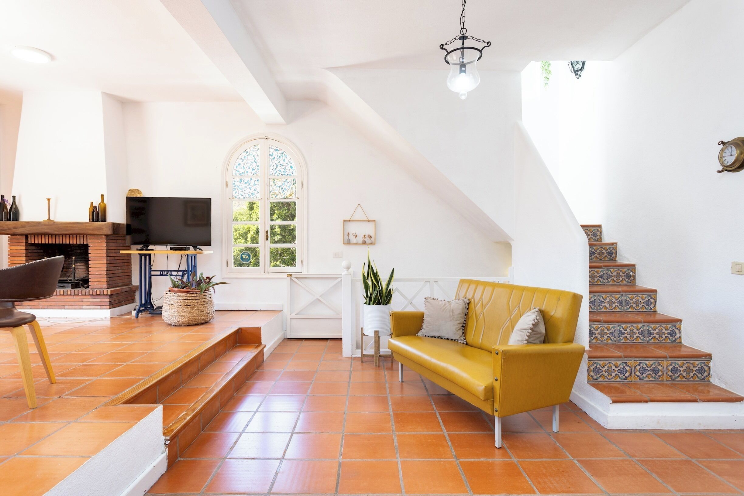 Ein geräumiger Wohnbereich mit Kamin, einem gelben Sofa, einem Fernseher und einer Treppe mit gemusterten Fliesen.