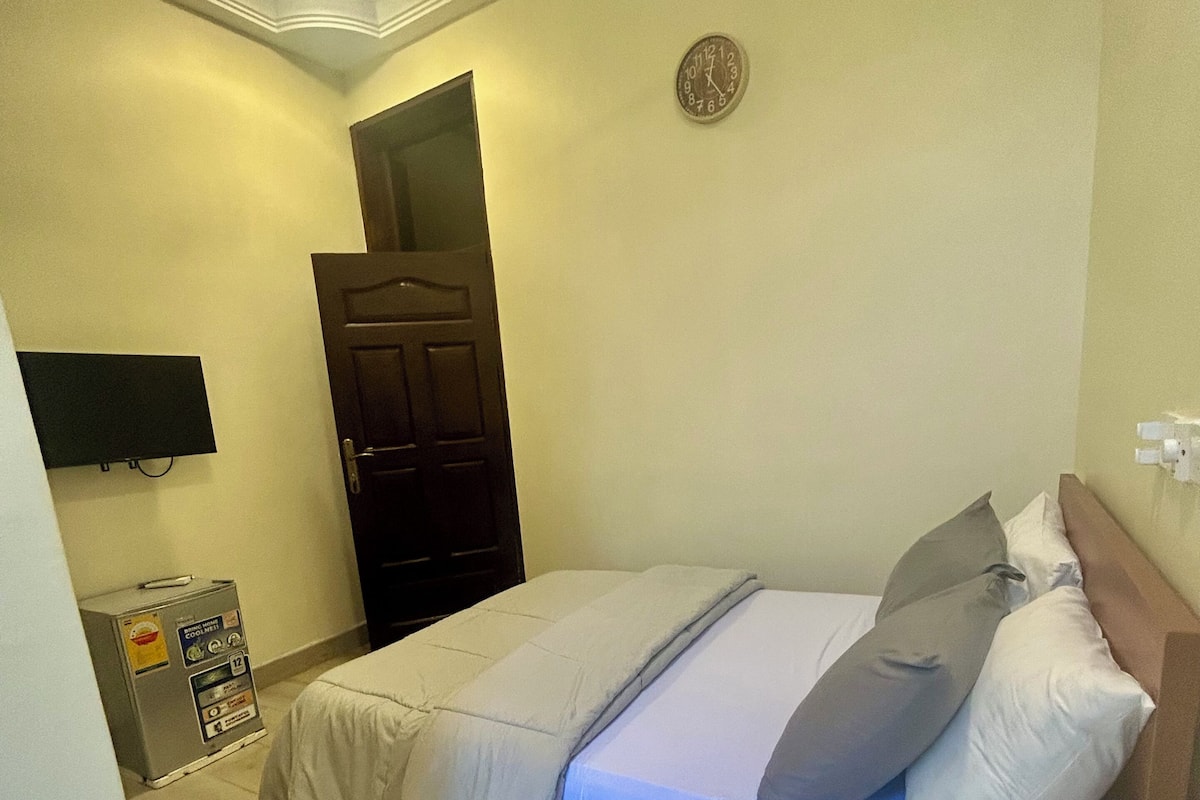 Un dormitorio con cama, almohadas, televisor, puerta y máquina expendedora.