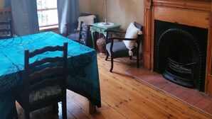 Um quarto com piso de madeira, uma mesa com toalha azul, uma cadeira, uma lareira e um quadro na parede.