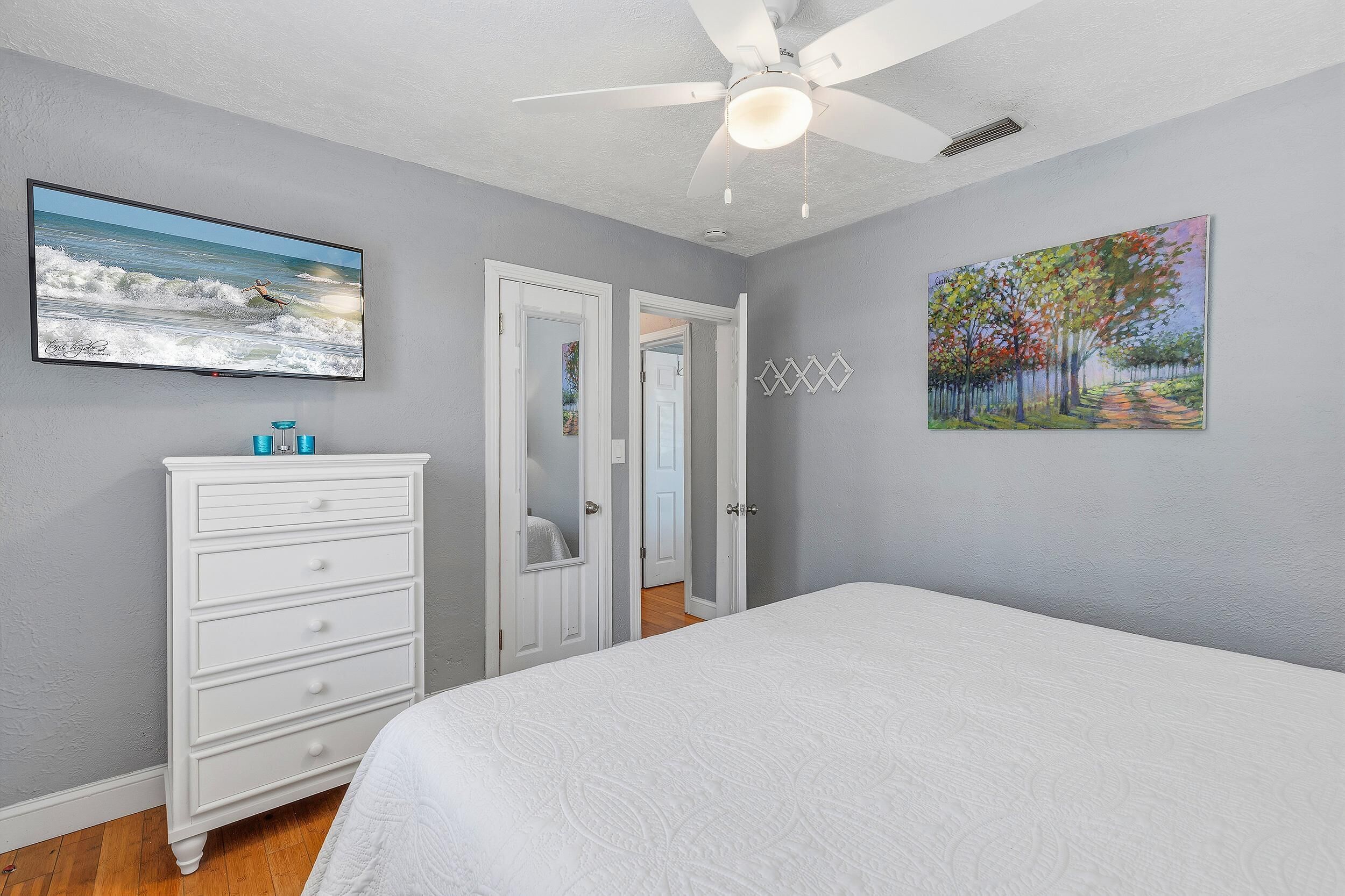 A bedroom with a bed, a dresser, a ceiling fan, a painting, and a television.