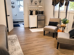 Un salon comprenant un canapé, des fauteuils, une table basse et un coin cuisine équipé d’appareils électroménagers.