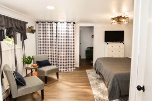 A bedroom with a bed, a chair, a desk, a TV, and a door leading to another room. - Northern Michigan Basecamp for Adventure (Onaway)