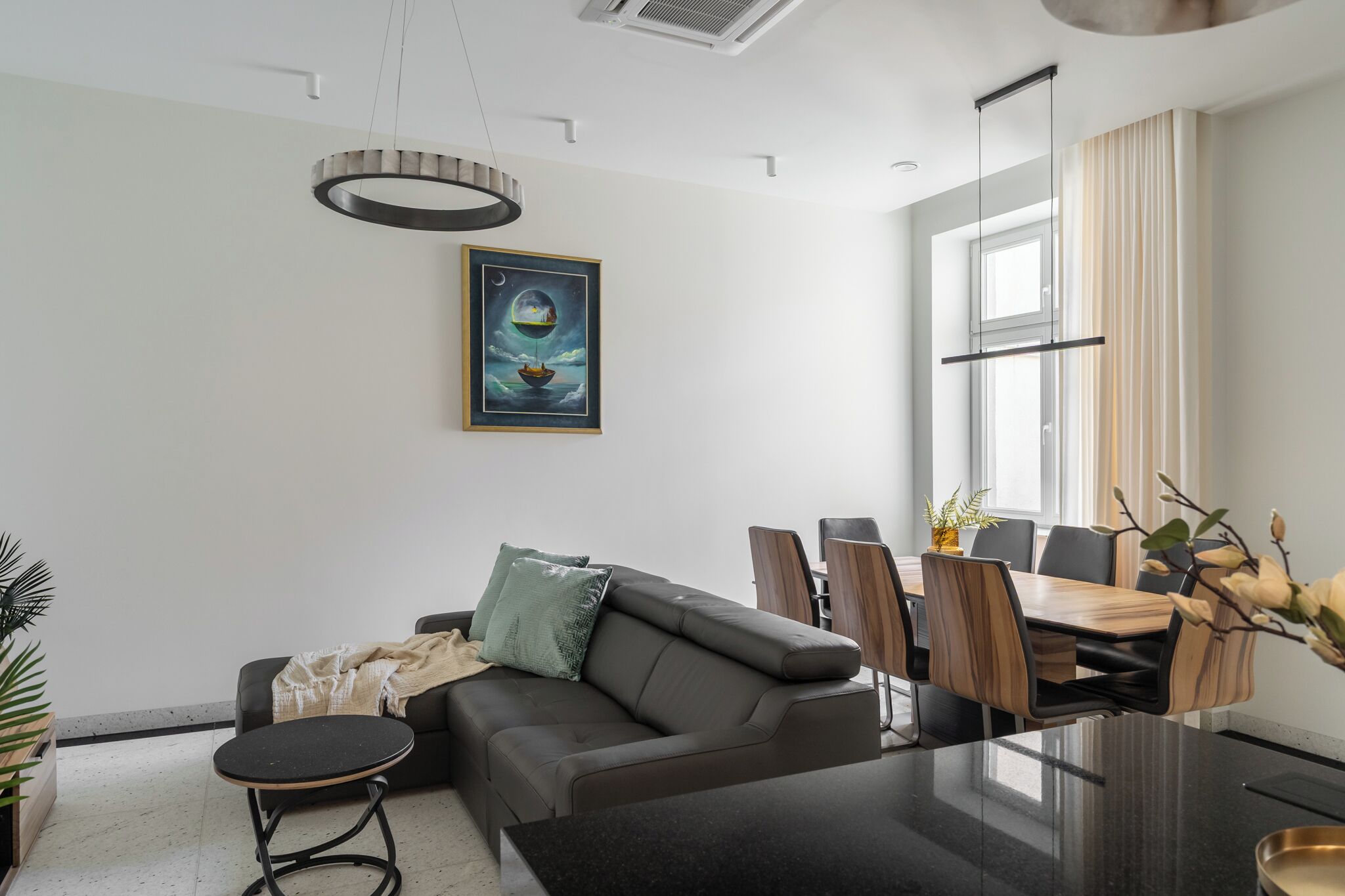 A modern living room with a sofa, a round coffee table, a dining table with chairs, a painting on the wall, and a large window.