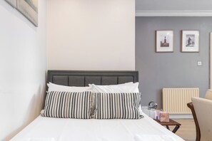 A neatly made bed with striped pillows, a grey headboard, and a bedside table with a lamp and a tissue box. - Chic Studio Retreat by Portobello Road (Greater London)