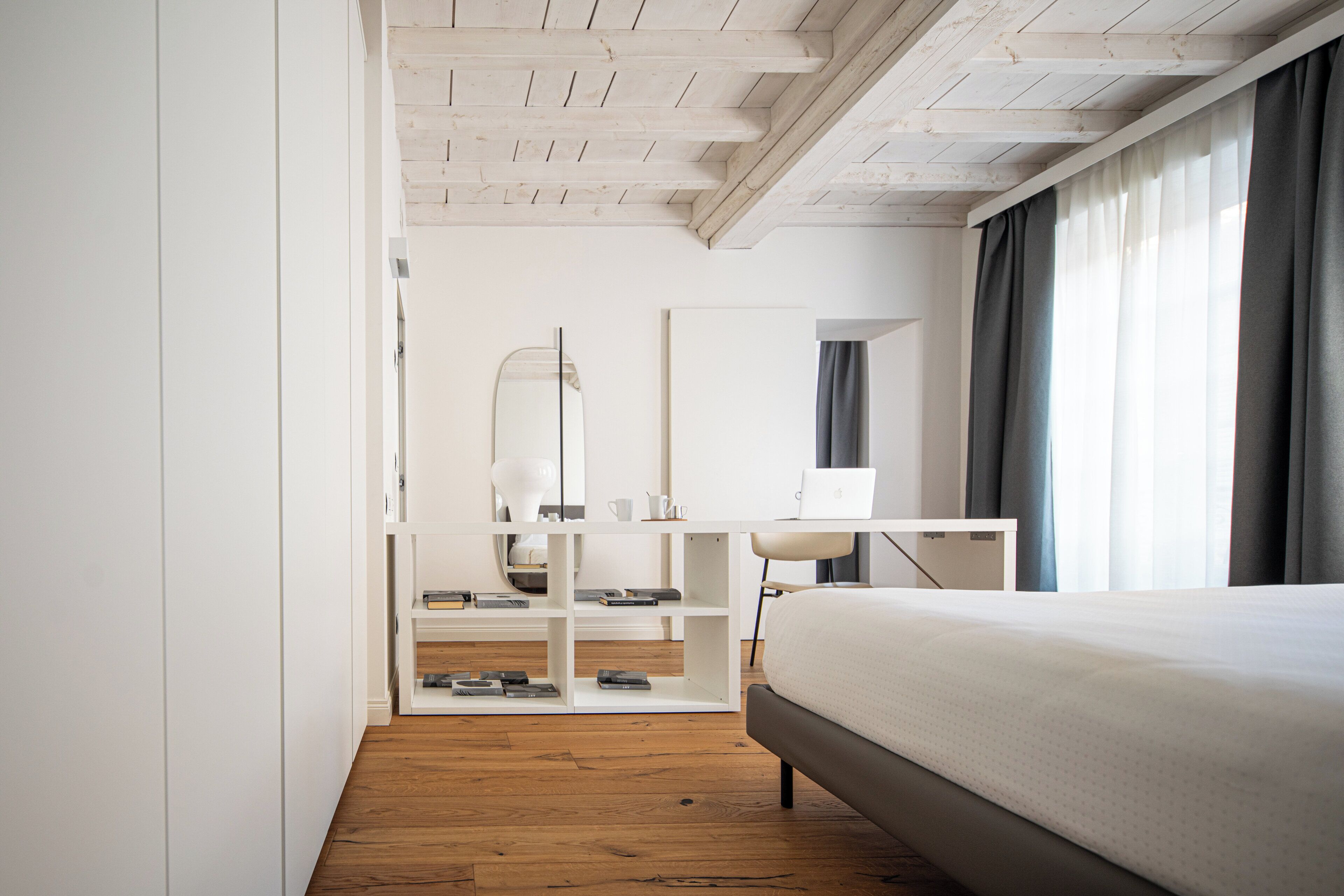A modern bedroom with a wooden floor, a bed, a desk with a laptop, and a large mirror.