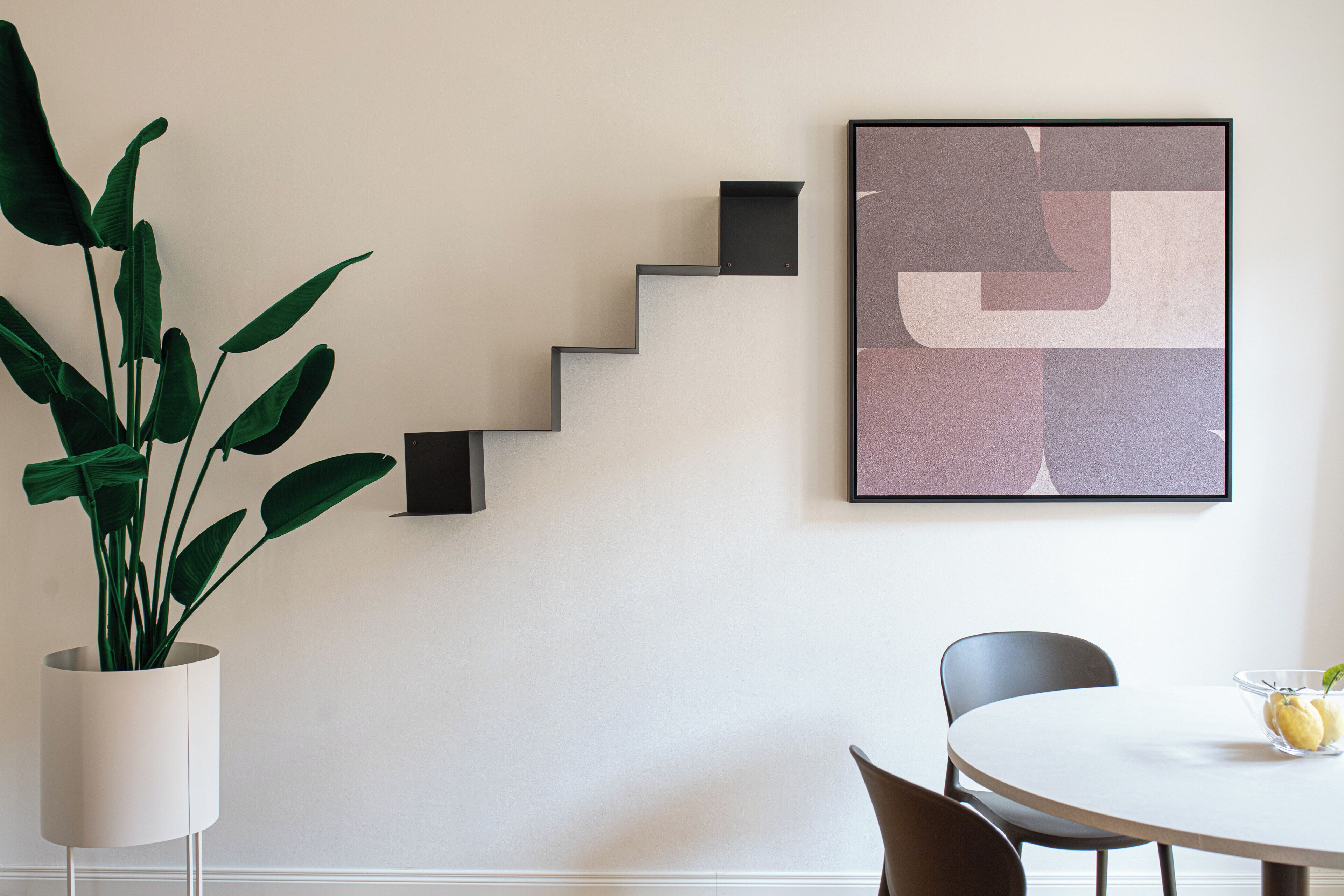 A modern interior with a staircase, a framed abstract artwork, a potted plant, and a dining area with chairs and a table.