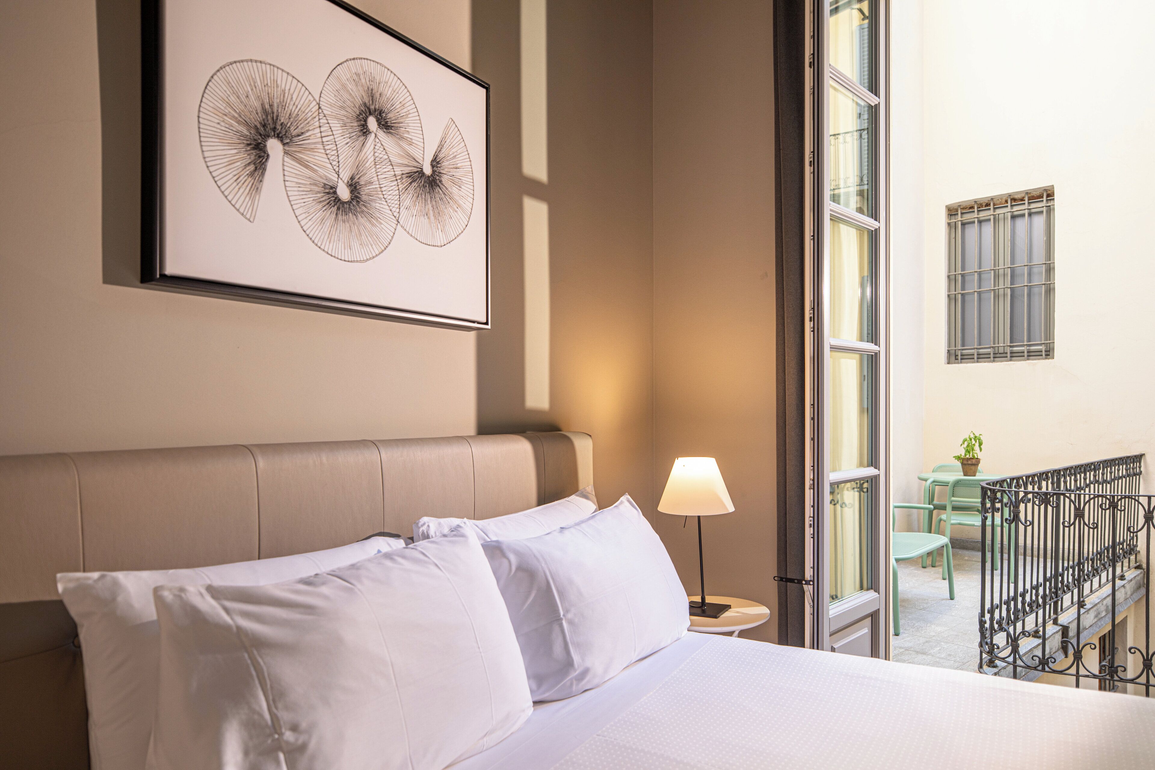 Habitación de hotel con cama, un cuadro enmarcado encima, ventana con vista al exterior y una pequeña mesa con lámpara.