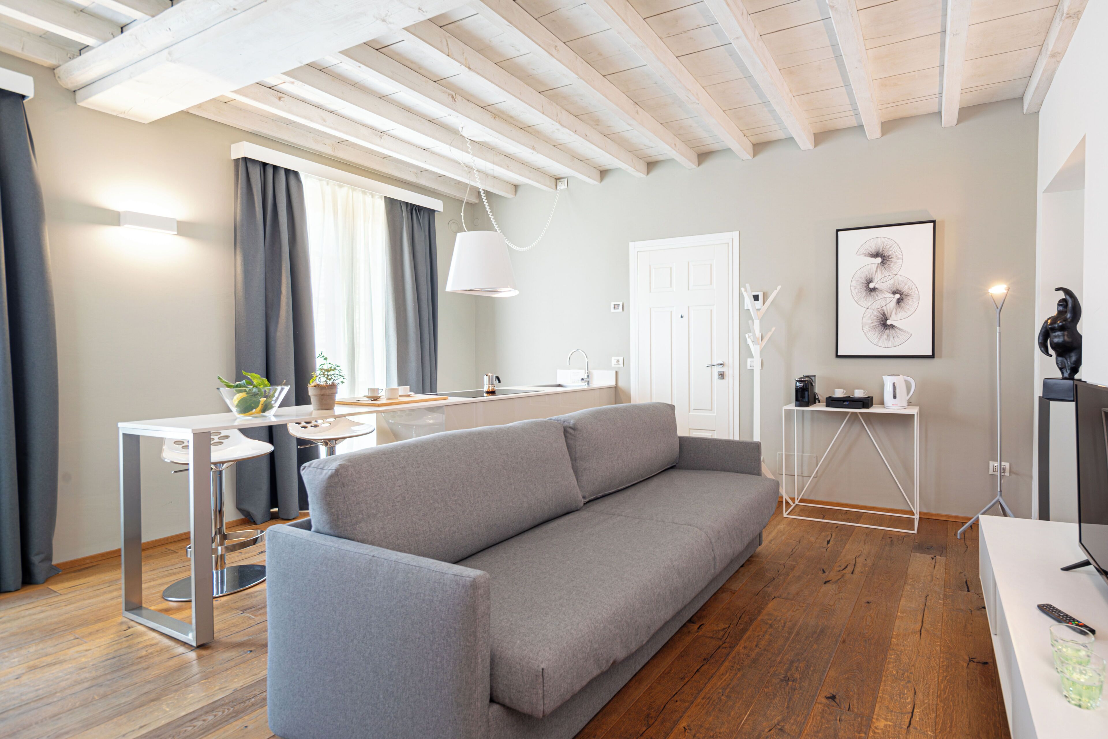 Una sala de estar moderna con un sofá gris, piso de madera, techo blanco y una zona de cocina al fondo.