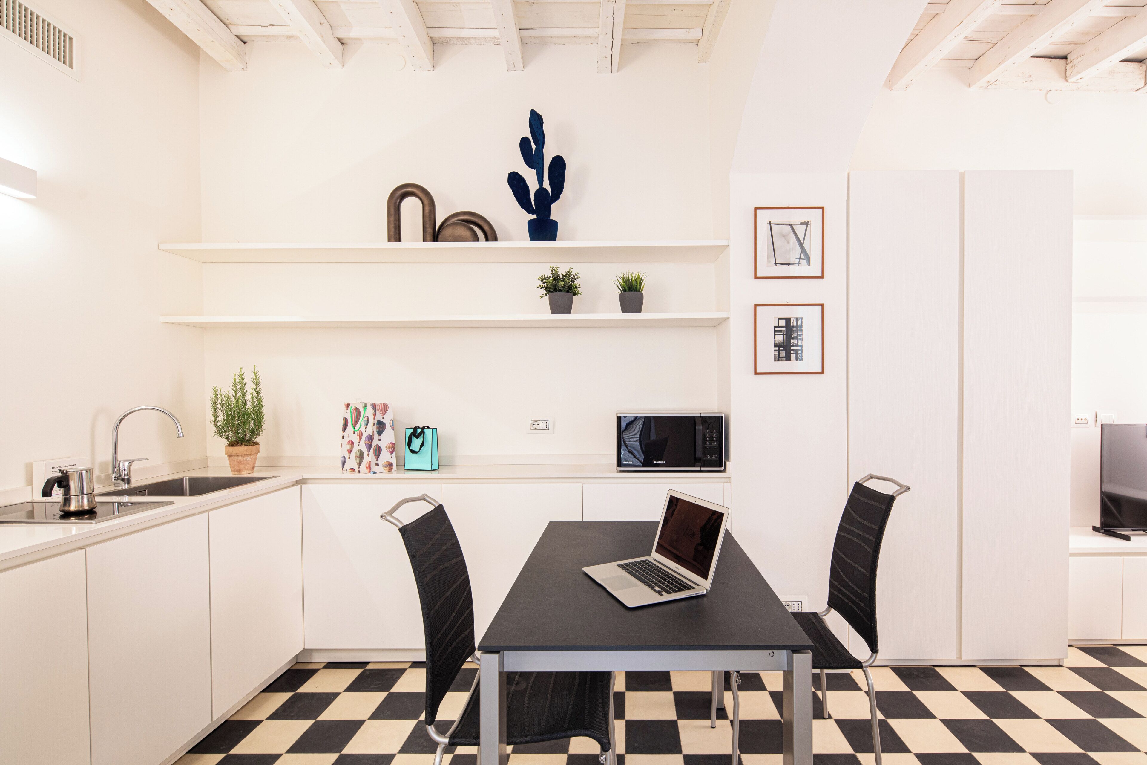 Une cuisine moderne avec des meubles blancs, un sol à carreaux noirs et blancs, une table à manger avec des chaises, un four à micro-ondes et un ordinateur portable posé sur la table.