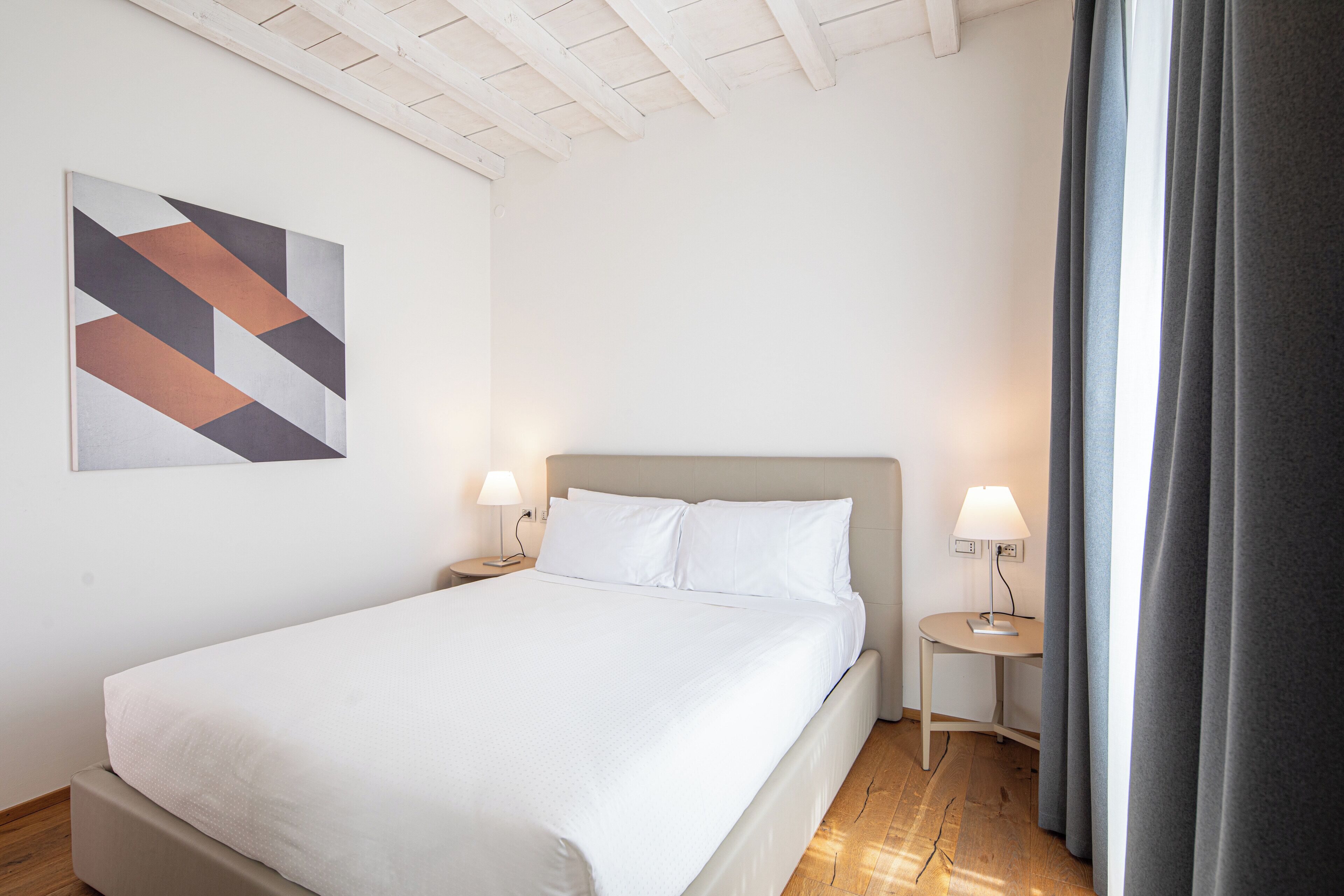 A hotel room with a bed, two bedside tables with lamps, a wall-mounted abstract painting, and a window with curtains.