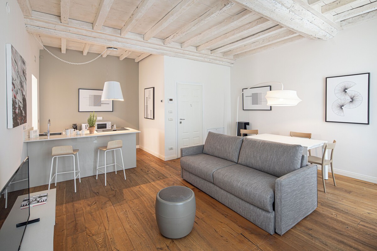 Un espacio habitable moderno con un sofá gris, piso de madera, una zona de comedor con una mesa blanca y una cocina con encimera blanca y taburetes de bar.