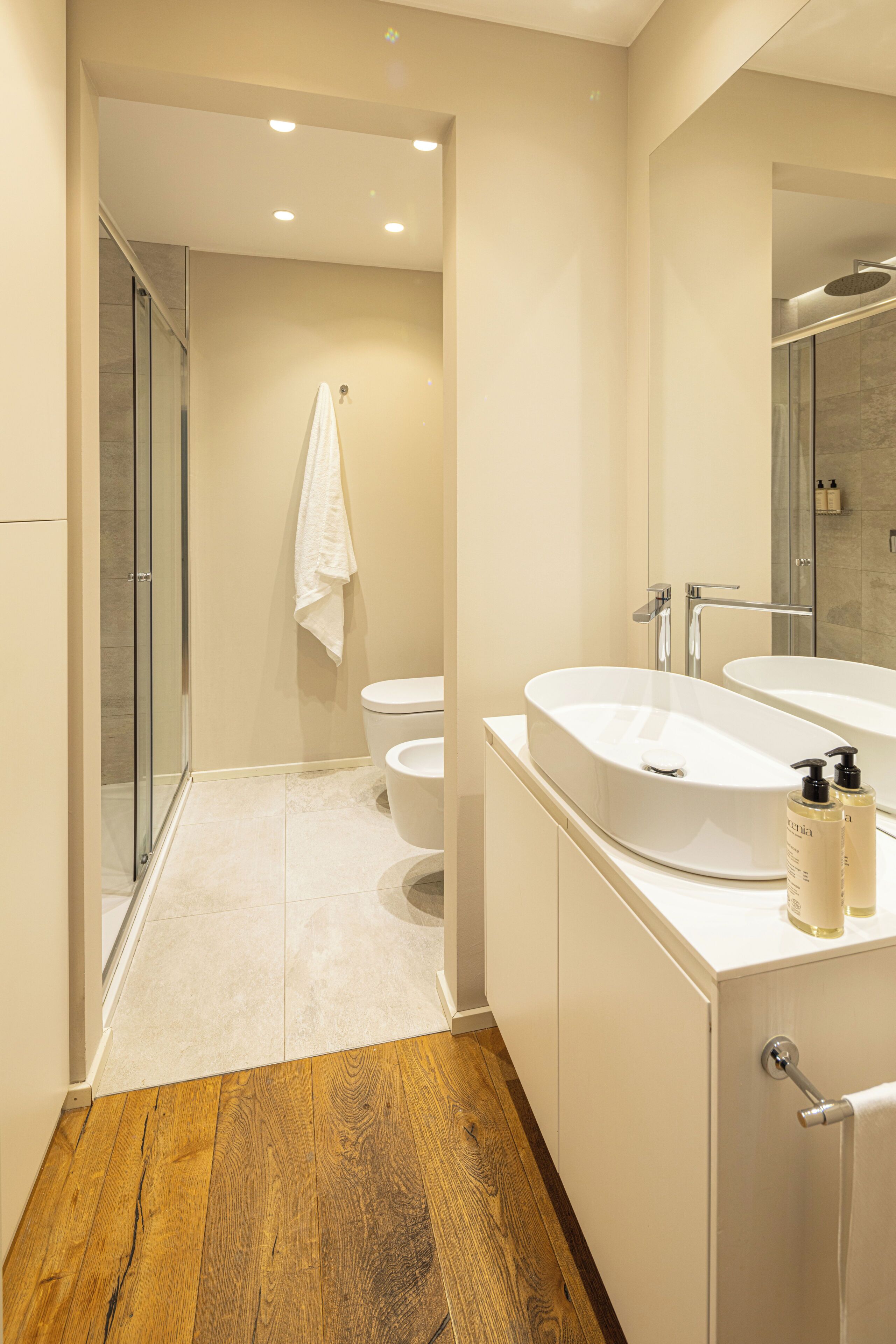 A modern bathroom with a glass shower enclosure, a white sink with a mirror, and a wooden floor.