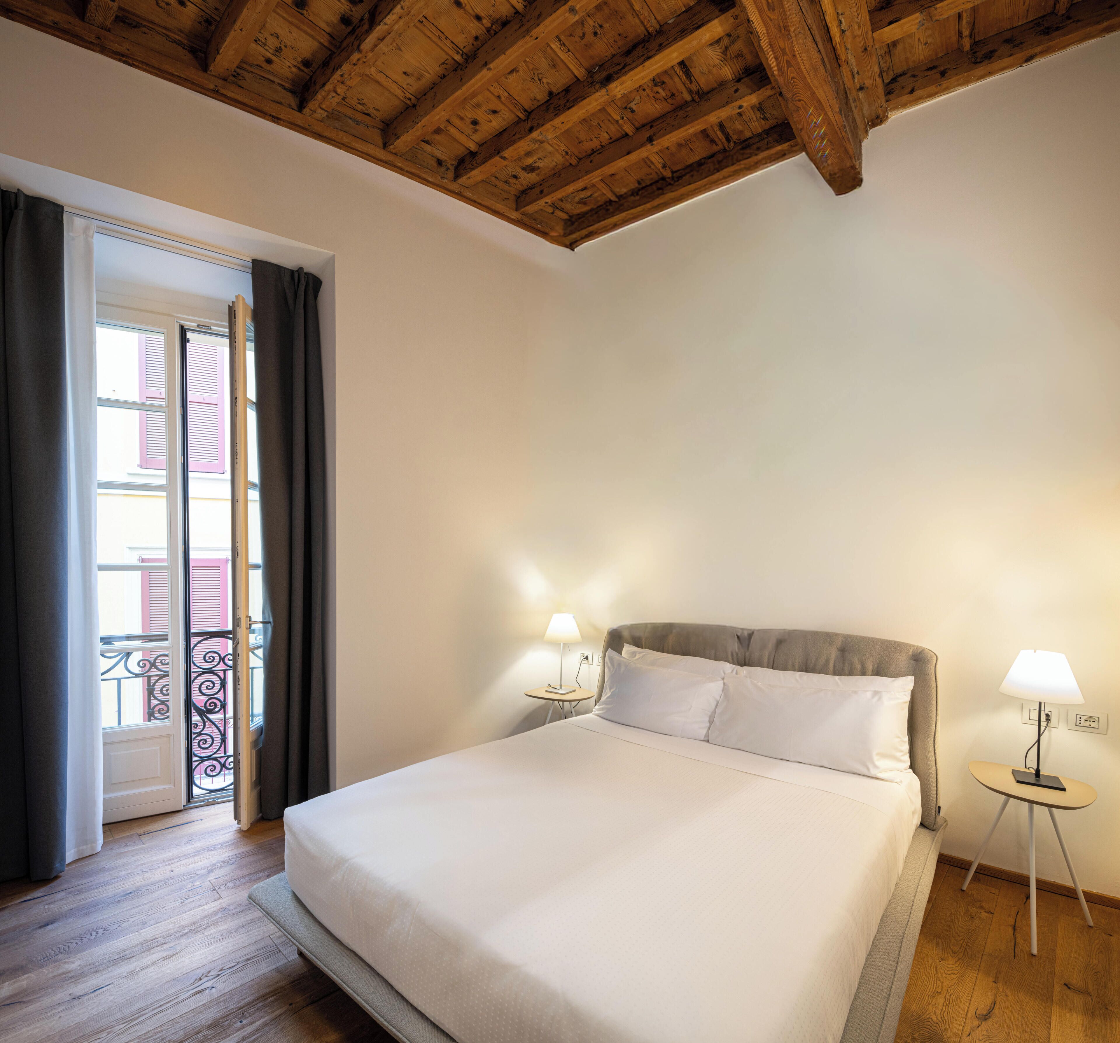 Habitación de hotel con techo de madera, cama con ropa de cama blanca, mesita de noche con lámpara y vista a un balcón con contraventanas rosas.