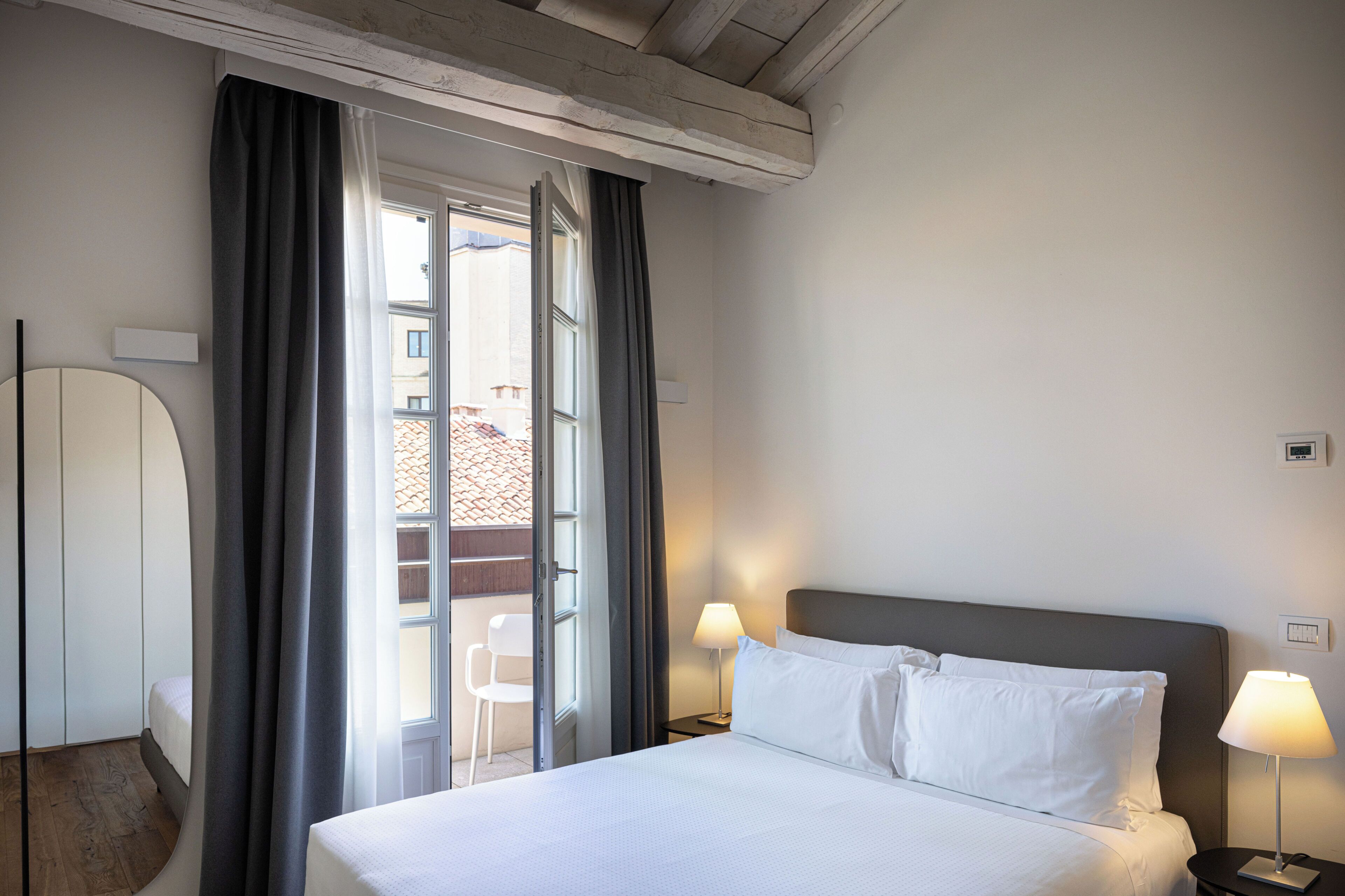 A hotel room with a bed, two bedside lamps, a window with curtains, a view of the outside, and a wall-mounted air conditioning unit.