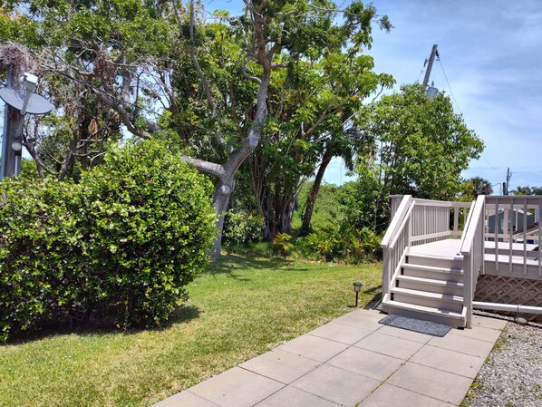Una propiedad residencial con camino de entrada pavimentado, escalera de madera y un jardín exuberante con árboles y arbustos maduros.