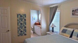 A bedroom with a bed, a nightstand, a lamp, a window with curtains, a door, and a wall with a picture. - Charming Sebastian Home Near the Waterfront Activities. (Sebastian)