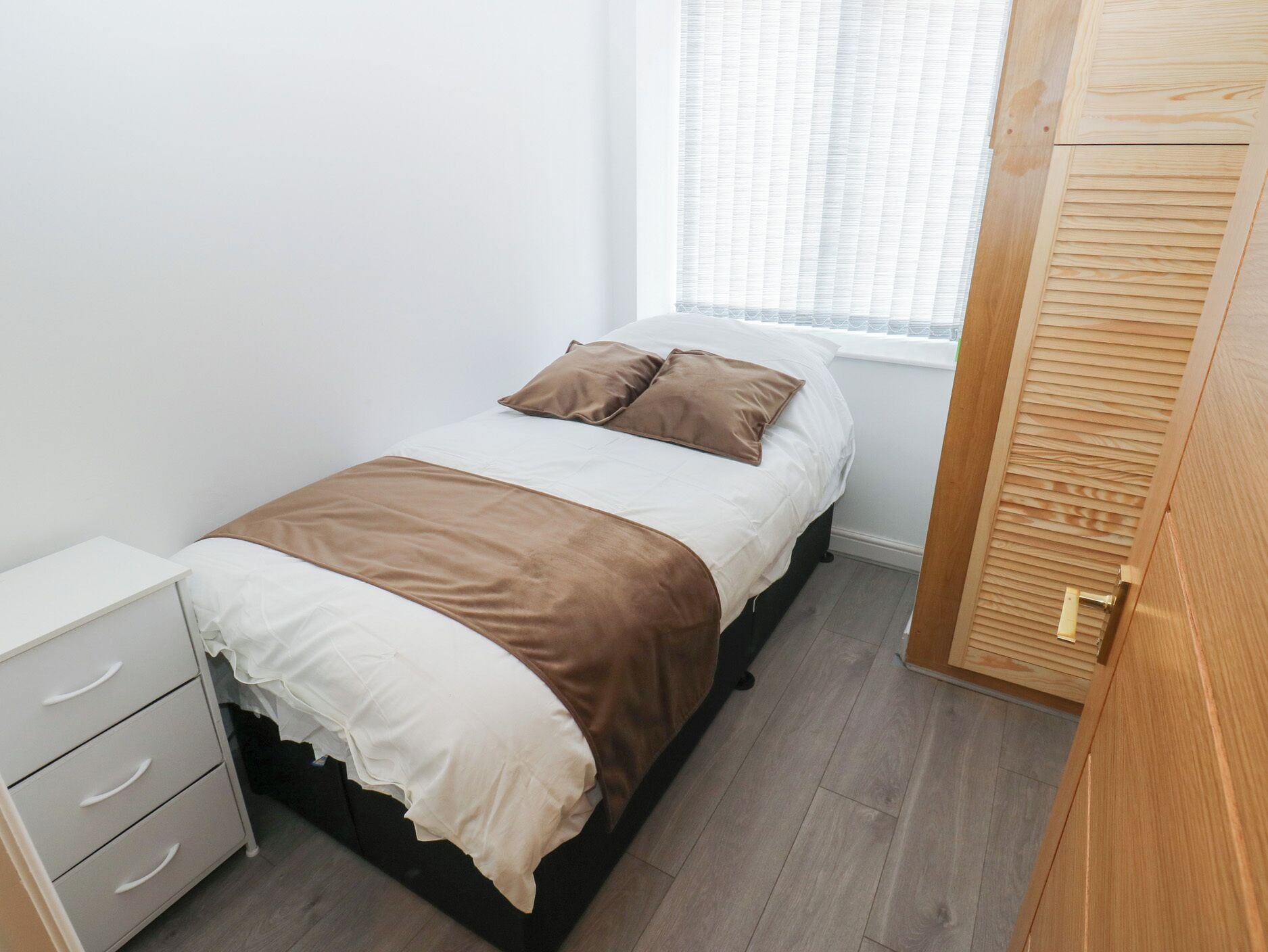 Un dormitorio con cama, mesita de noche con cajones, ventana con persianas y armario de madera.