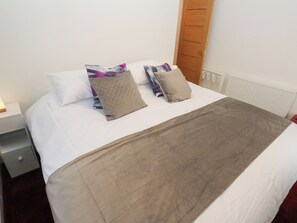 Una cama bien hecha con edredón blanco y dos almohadas, una manta marrón, una mesita de noche con lámpara y una puerta de madera.