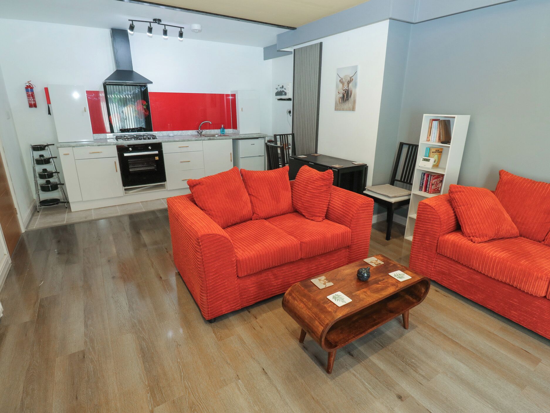 Un espace de vie compact comprenant un coin cuisine, deux canapés rouges, une table basse en bois et une bibliothèque.