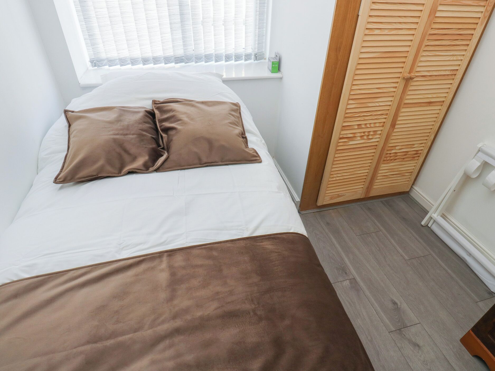 Una cama con ropa de cama blanca y marrón, una ventana con persianas, un armario de madera y un radiador blanco.