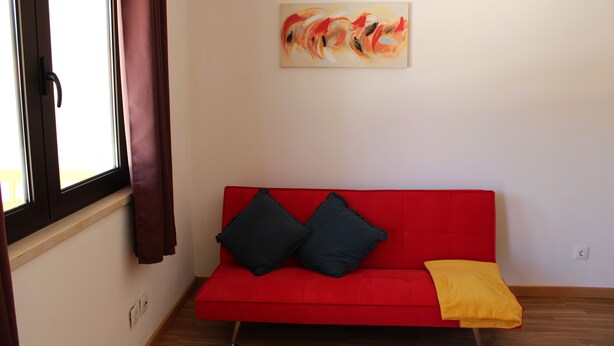 Un sofá rojo con dos cojines negros y una manta amarilla, un cuadro en la pared y una ventana con cortinas.