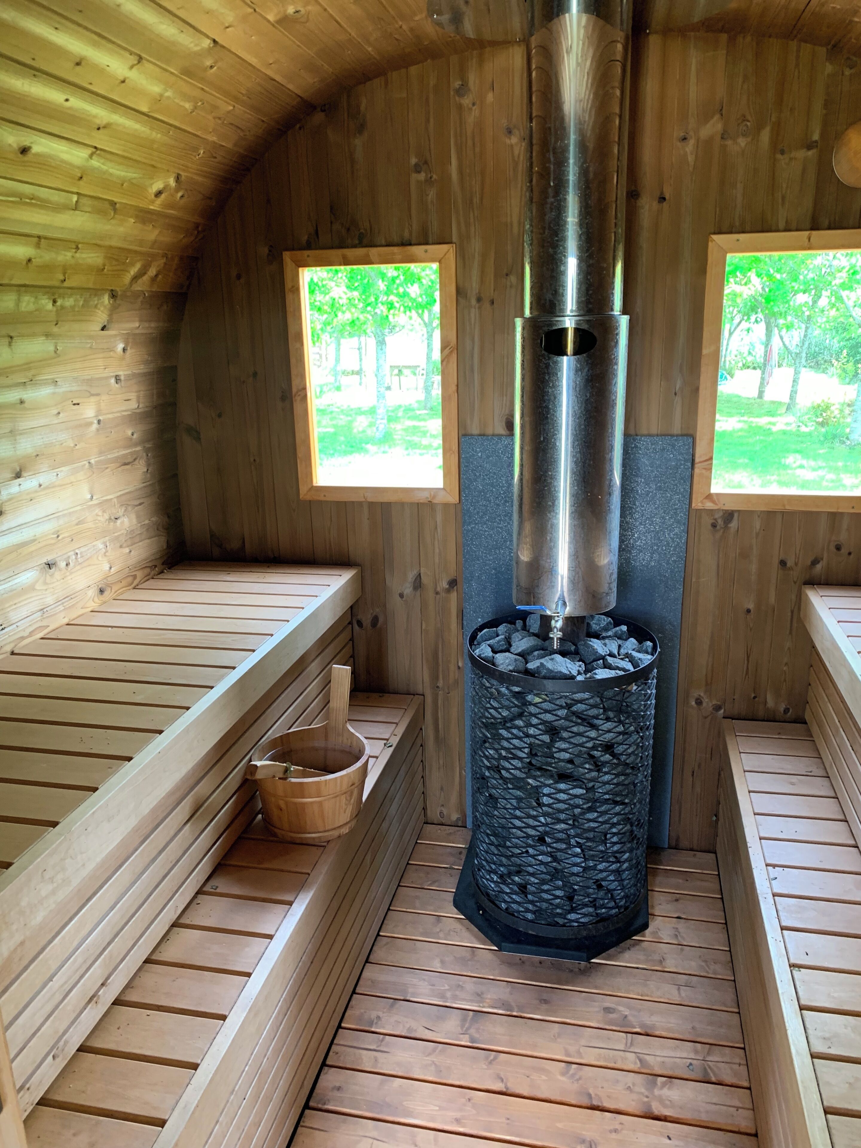 Un sauna con bancos de madera, un calentador de piedra central y dos ventanas con vistas al follaje.