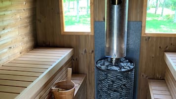 Un sauna con bancos de madera, un calentador de piedra central y dos ventanas con vistas al follaje.