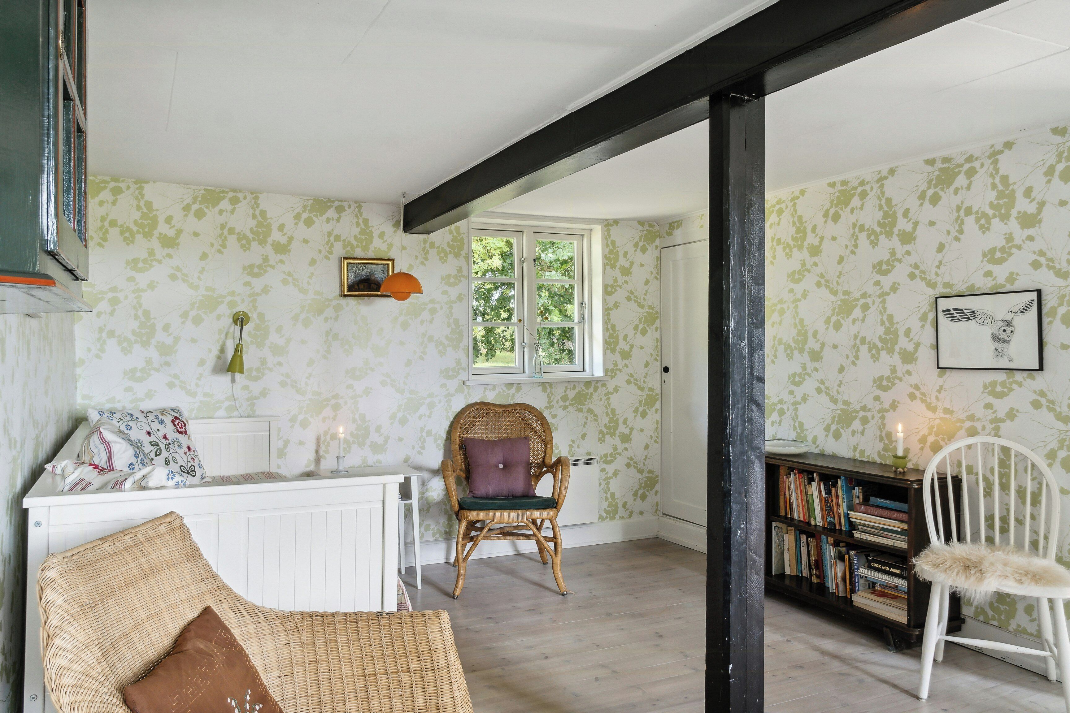A room with a white radiator, a wicker chair with a cushion, a white chair, a bookshelf, and a window with a view of trees.